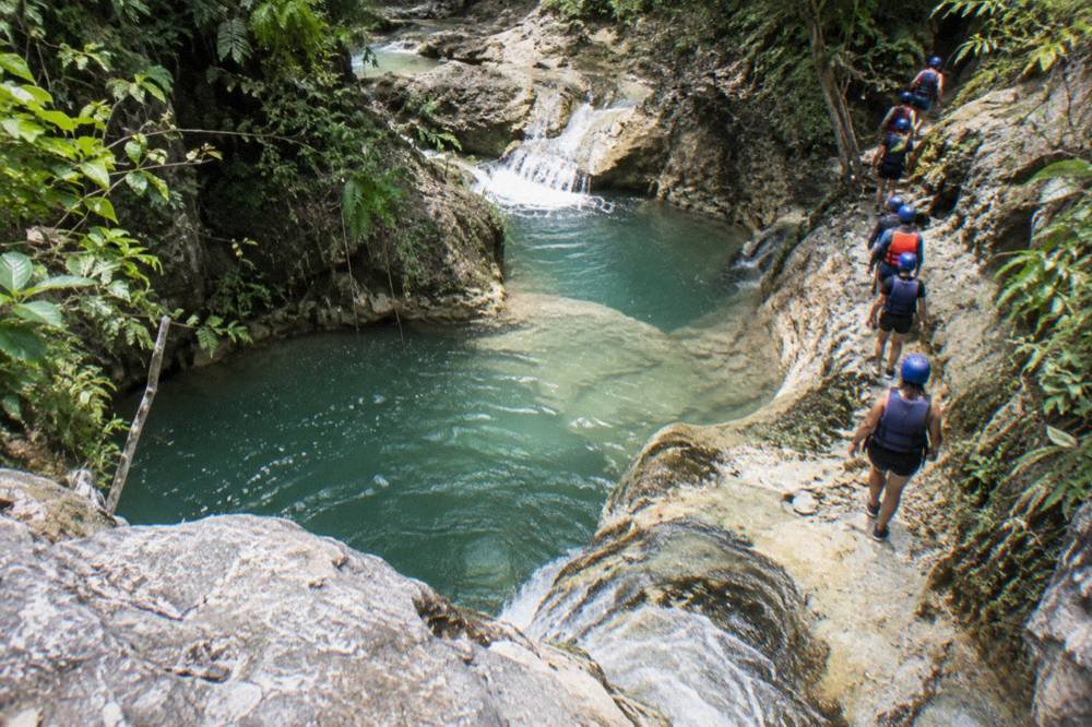 Visitors in helmets and life jackets walking single file along a rocky riverbank between pools