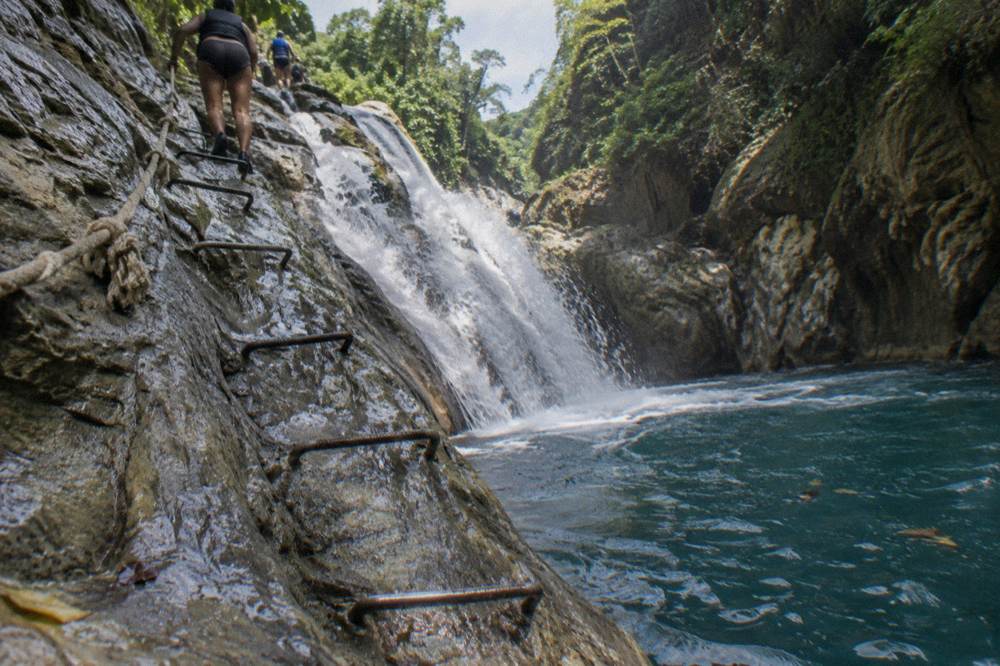 Wooden ladder bolted to a canyon wall beside a sliding waterfall into a deep blue pool