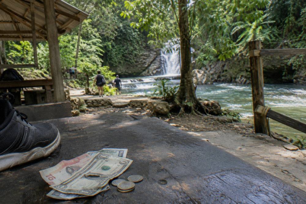 Weathered peso and dollar banknotes held in a hand beside a hiking shoe on a wooden bench