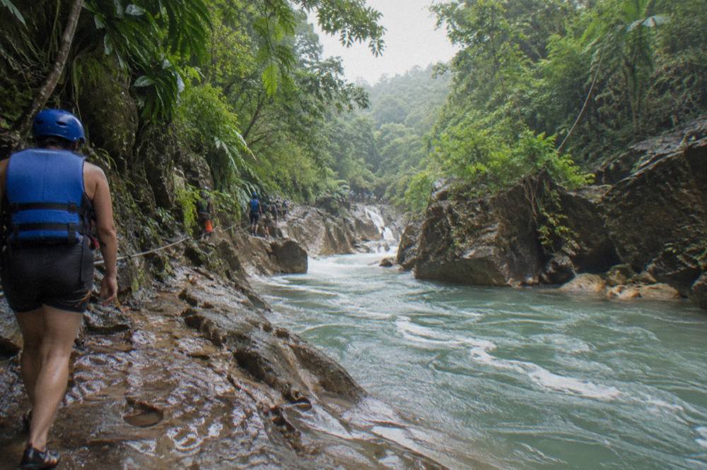 A hiker in helmet and life jacket walking along a wet rocky path beside a flowing river in a forested canyon
