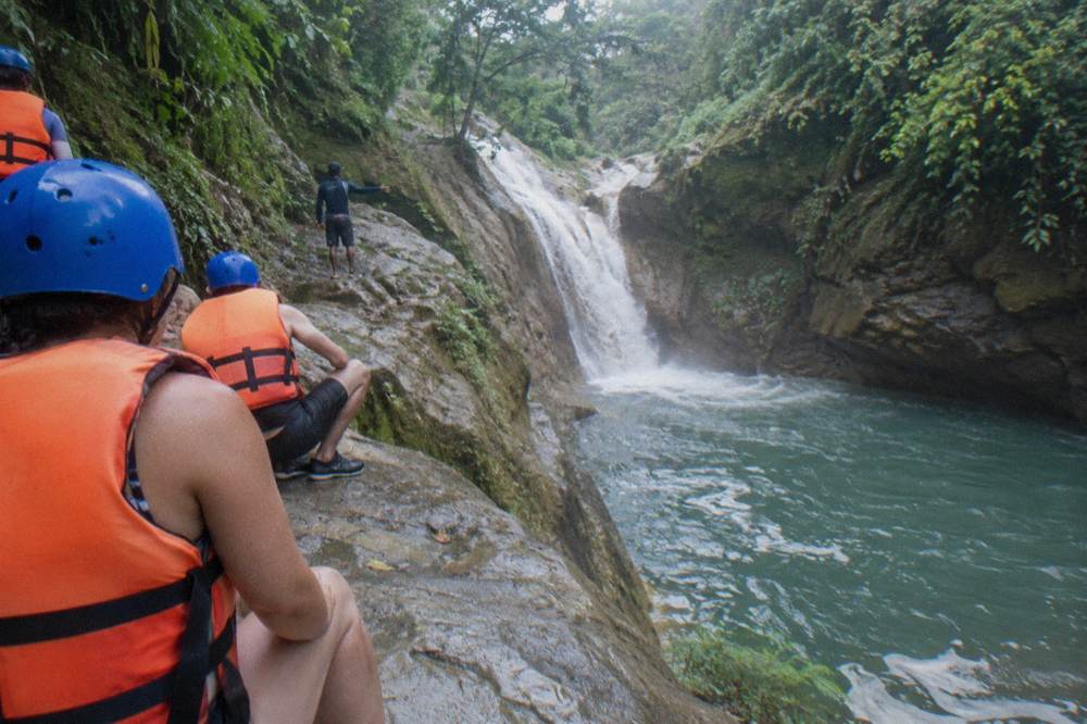 A group of visitors in helmets and life jackets waiting on rocks beside a waterfall pool while a guide gestures instructions