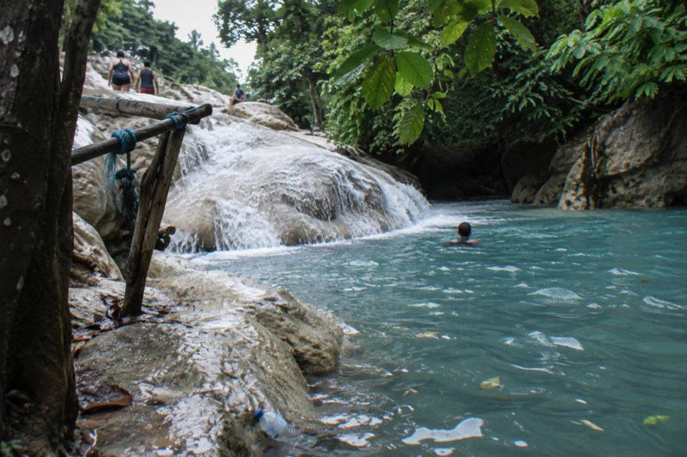 Water flowing over smooth limestone ledges into a deep blue pool framed by green trees