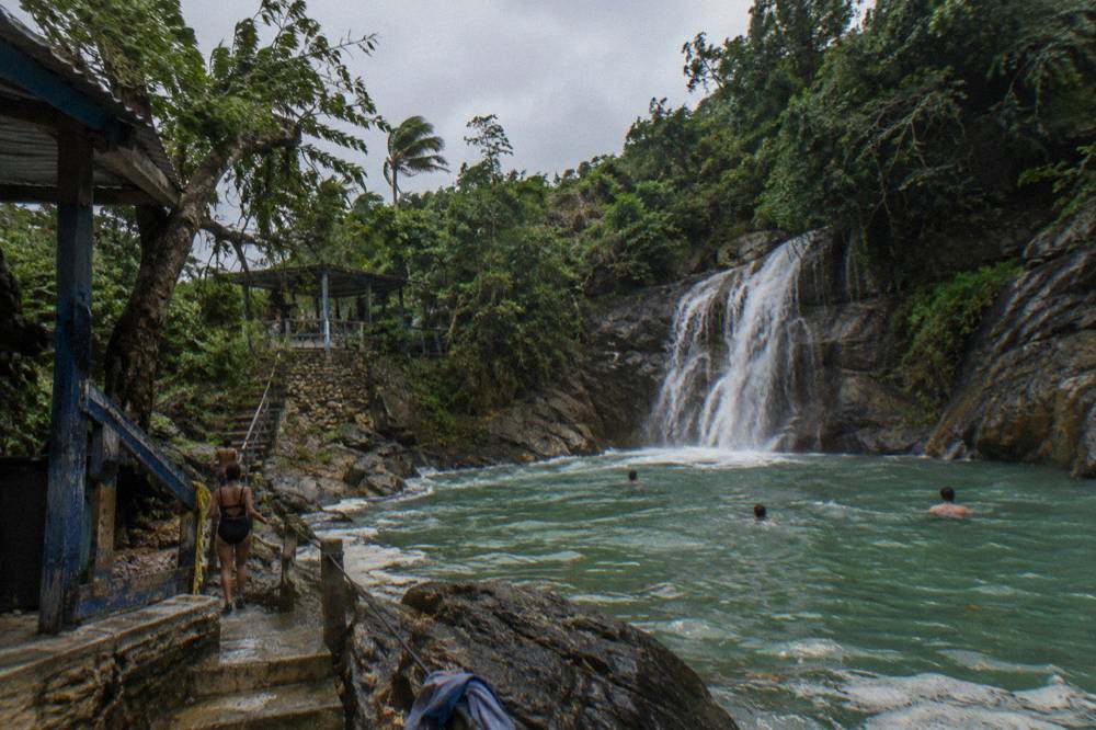 Waterfall pouring over dark rock into a wide green pool surrounded by tropical trees