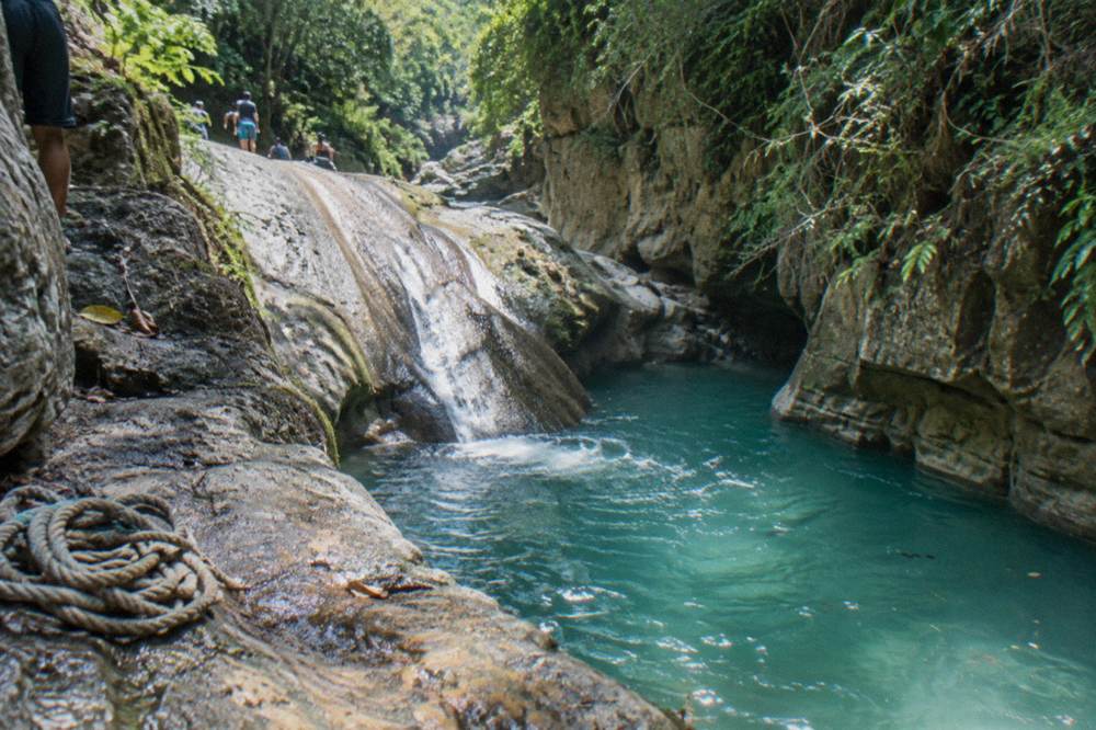 Calm turquoise pool at the base of a rock slide in a green canyon