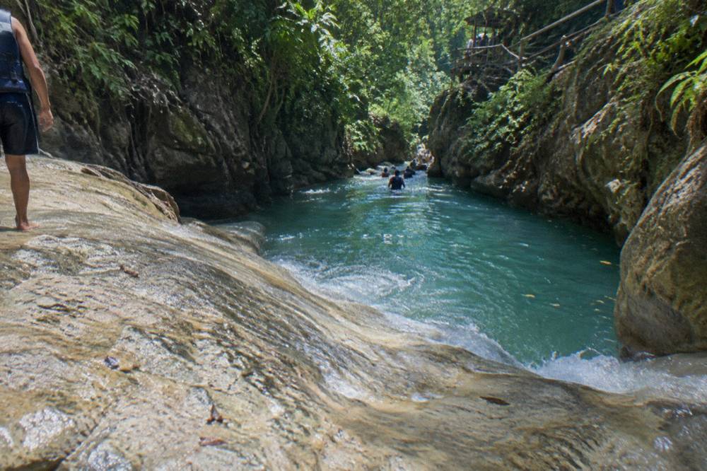 Turquoise water pooling at the base of a rock slide inside a narrow green canyon