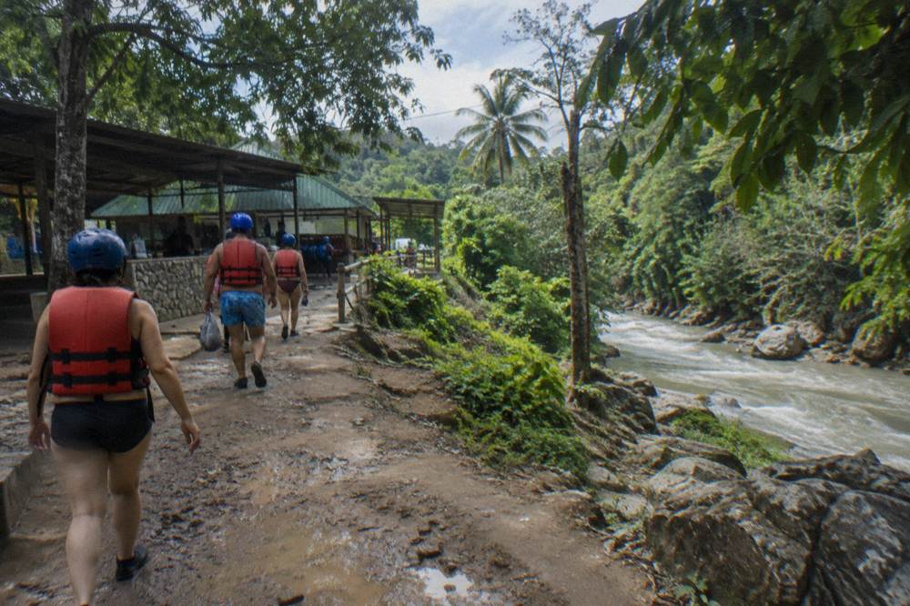 Visitors in helmets and life jackets walking a dirt trail through dense tropical forest beside a river