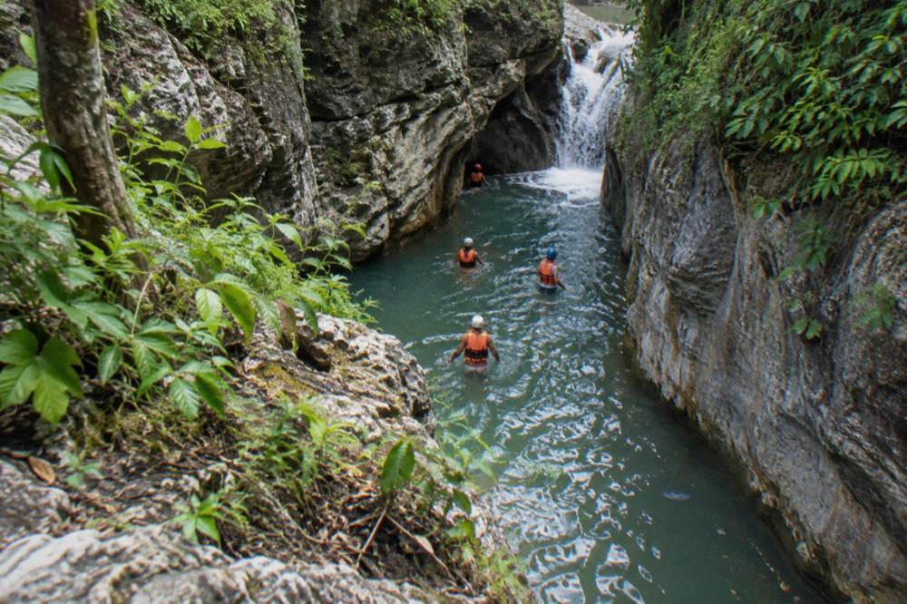 Tour group in helmets and life vests wading through a narrow canyon pool