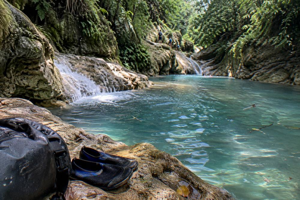 Dry bag and water shoes resting on a rock beside a calm turquoise pool