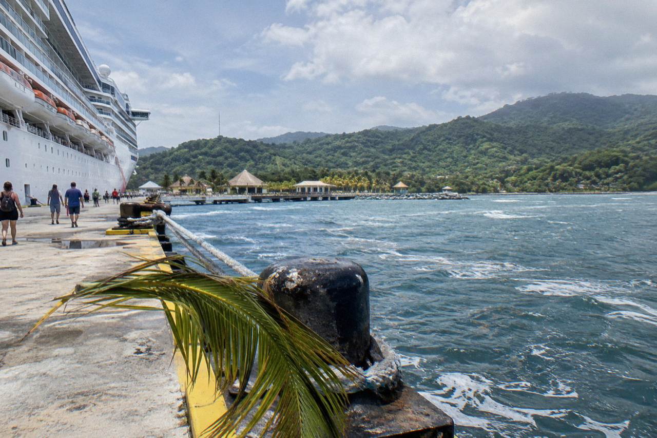 Cruise ship docked at a tropical port terminal with green hills behind the pier
