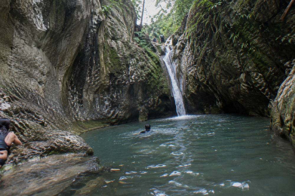Narrow canyon walls rising above a green pool with a small waterfall