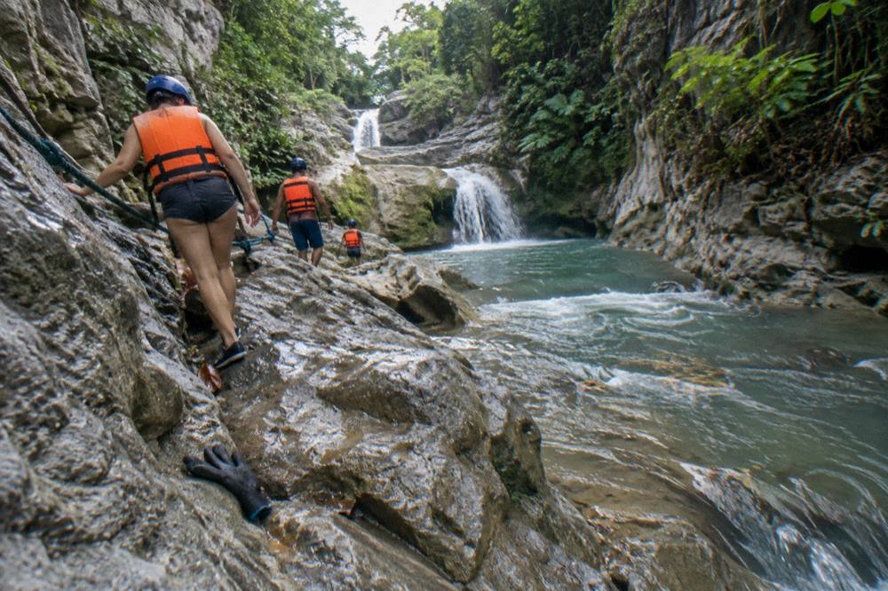 Small group in helmets and life jackets walking along a riverbank in a canyon