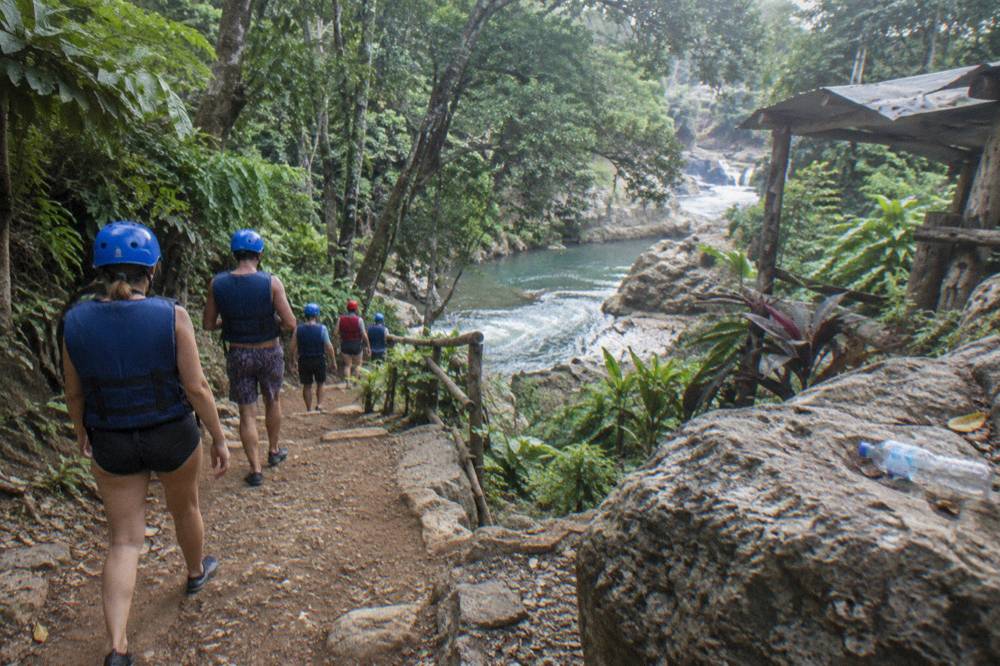 Visitors in helmets and life vests walking a dirt trail through dense green forest toward the river