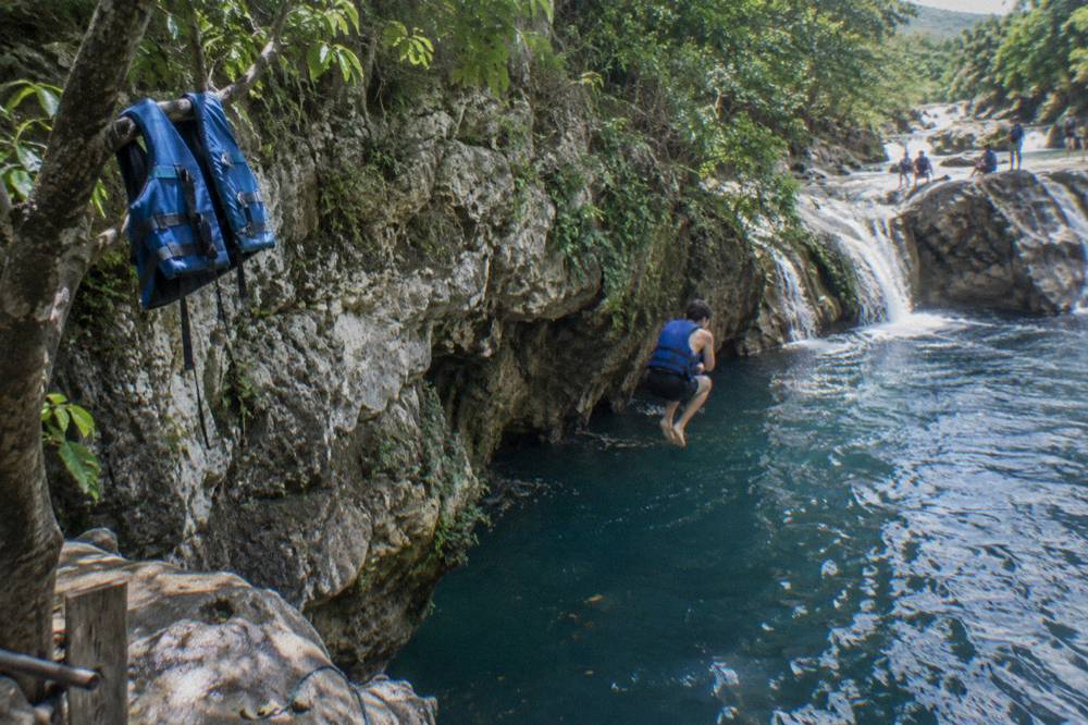 Swimmer in a life vest mid-air jumping from a rock ledge into a deep blue pool