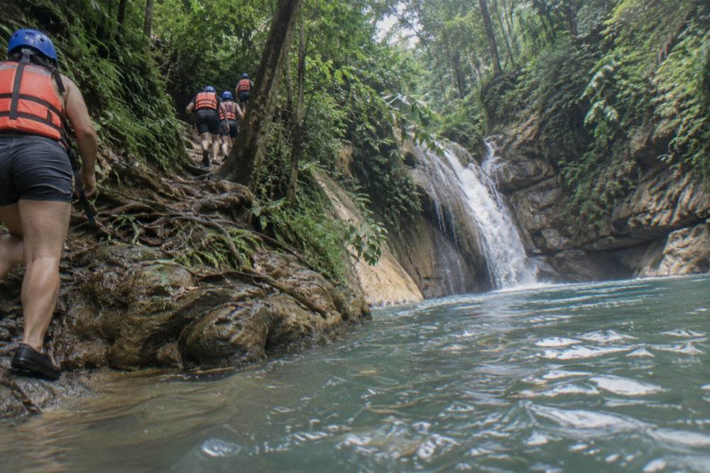 Hikers in helmets and life jackets walking up a jungle trail beside a river