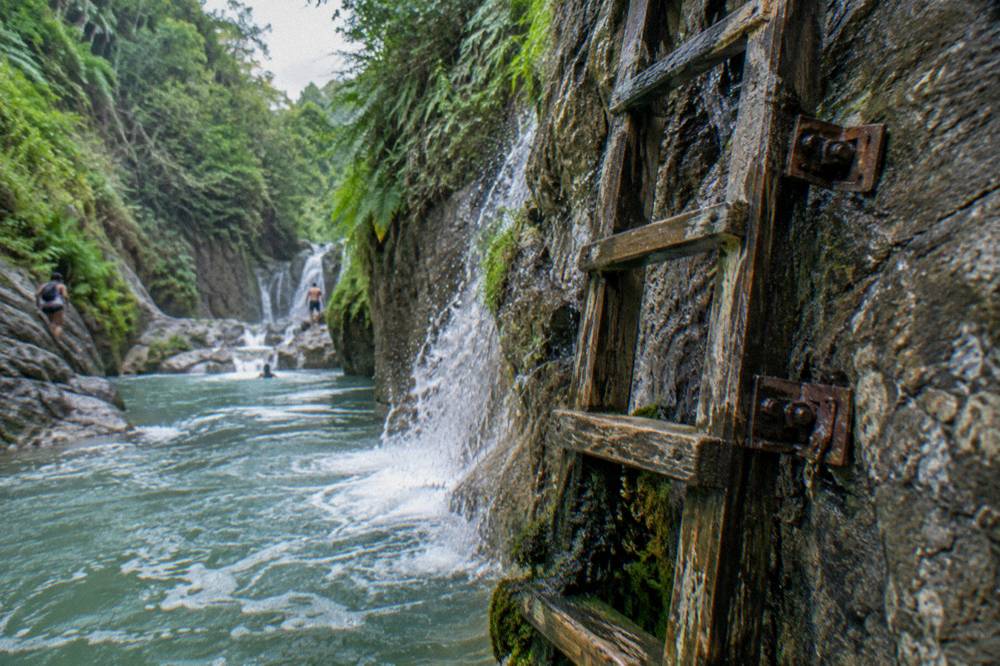 Wooden ladder and rock wall beside a cascading waterfall in a green canyon