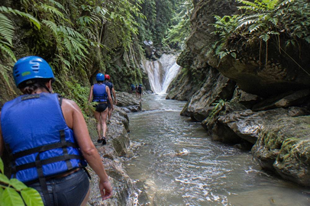 Hikers in helmets and life vests walking a narrow canyon trail beside a stream