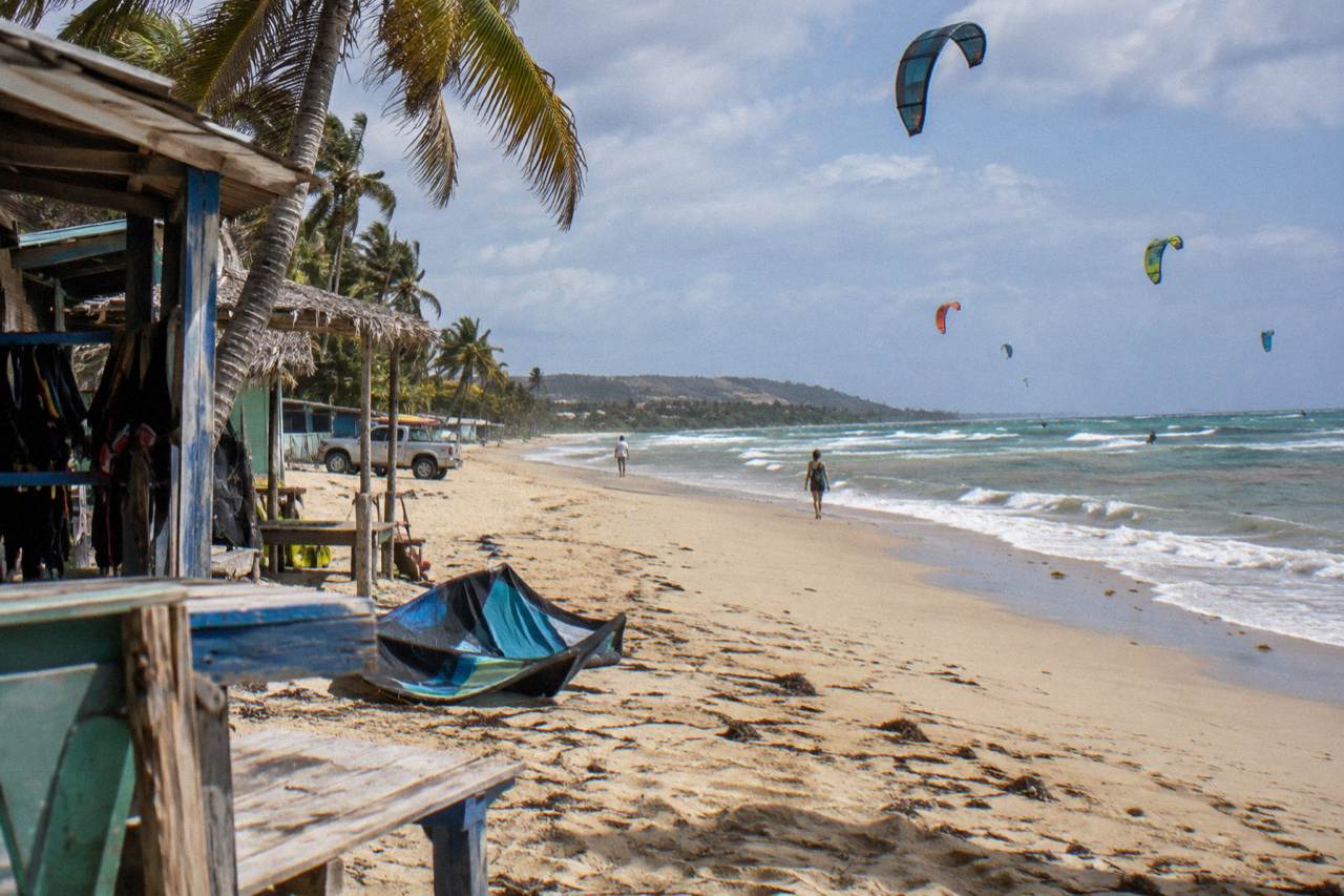 Cabarete beach with golden sand, kitesurf kites over a turquoise sea, palms and wooden shacks