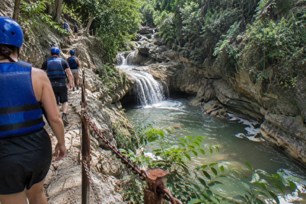 Hikers in helmets and life jackets walking a narrow trail above a river gorge