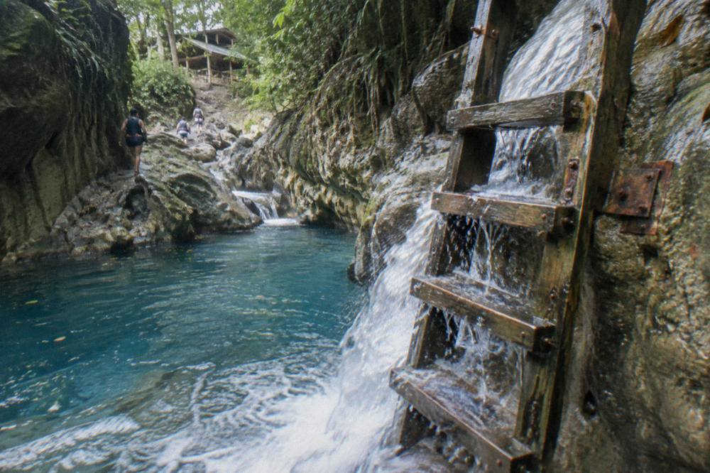 Small wooden ladder descending beside a waterfall into a clear blue pool