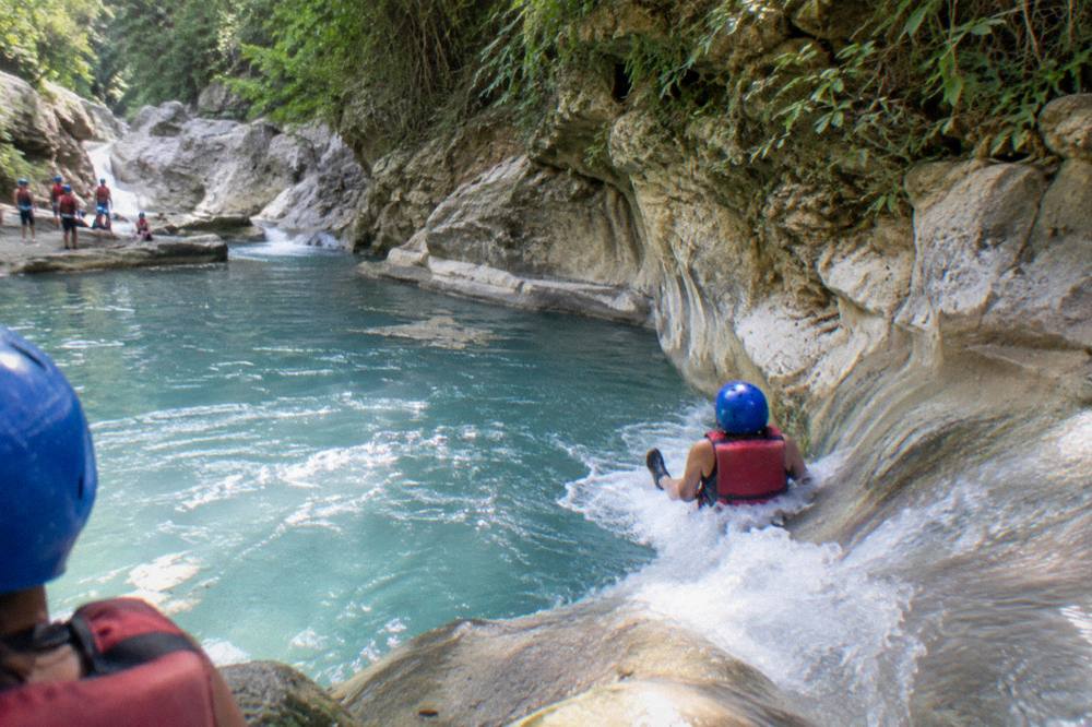 Visitors in helmets and life jackets sliding down a rock slide into a wide turquoise pool