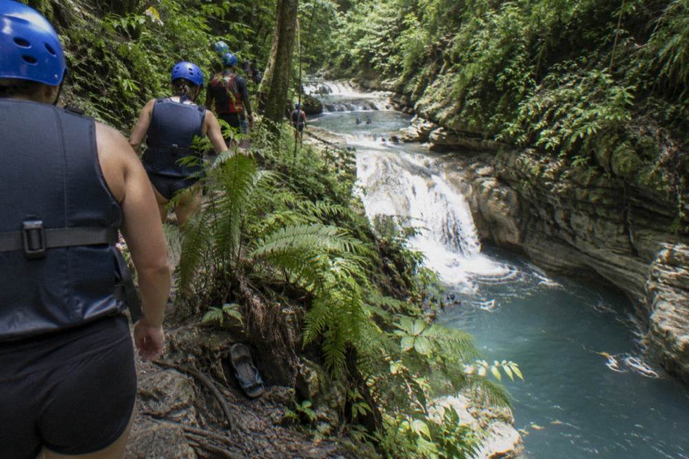 Hikers in helmets and life vests walking a narrow dirt trail above a river gorge