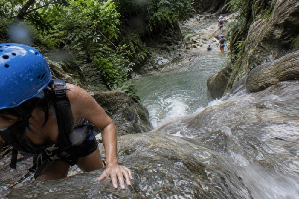 Hiker in a helmet gripping wet rock beside a sliding cascade
