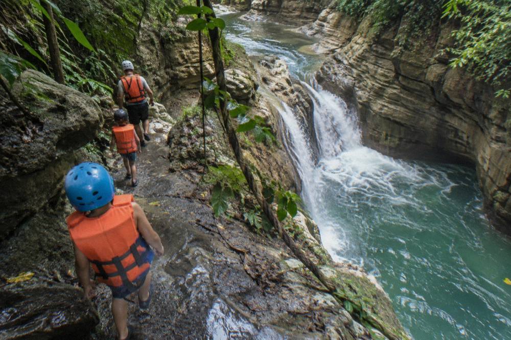 Family in helmets and life jackets walking a canyon trail beside a waterfall