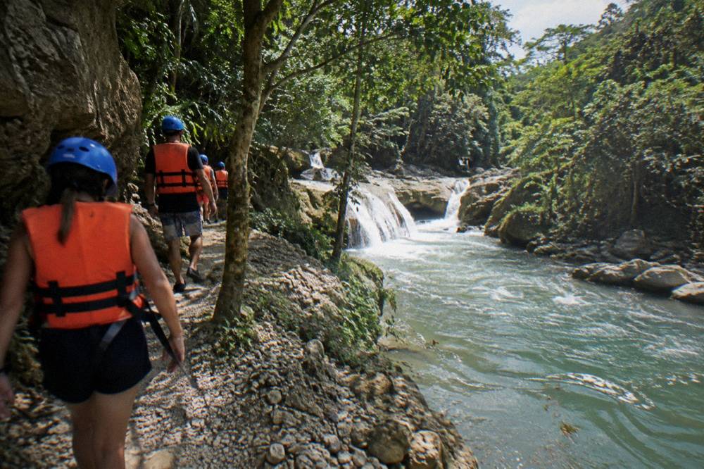 Visitors in helmets and life jackets walking a canyon trail beside a river
