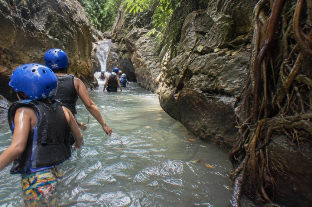 Family group in helmets and vests wading through a shallow river between rock walls
