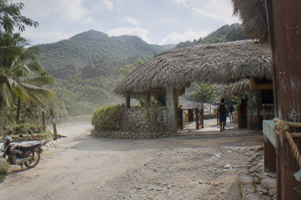 Roadside entrance area with wooden signage and green hills behind it