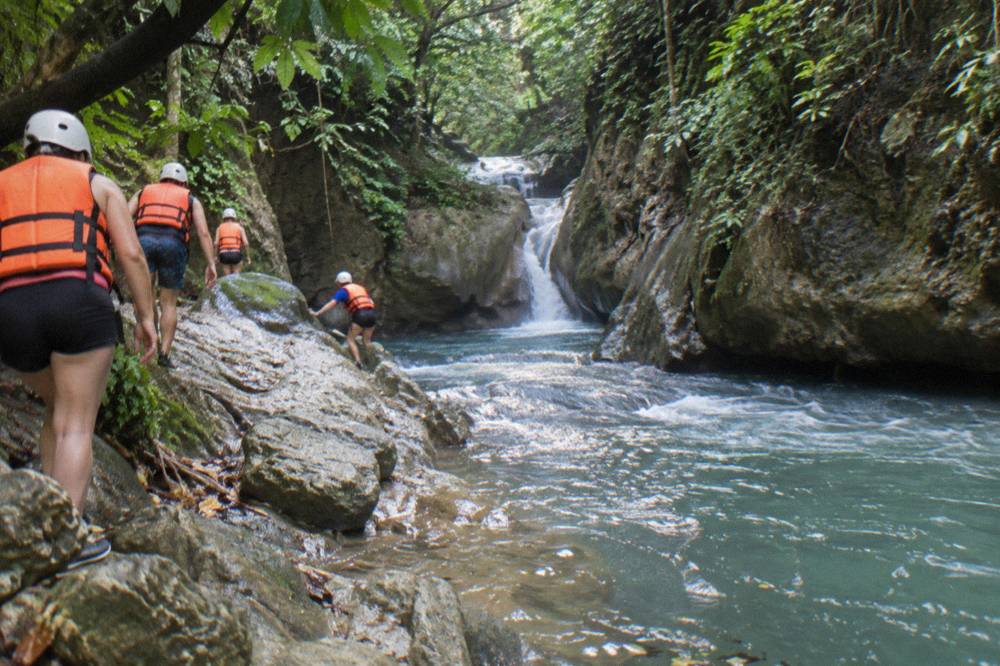 Small group in helmets and life jackets walking along a riverbank in a narrow green canyon