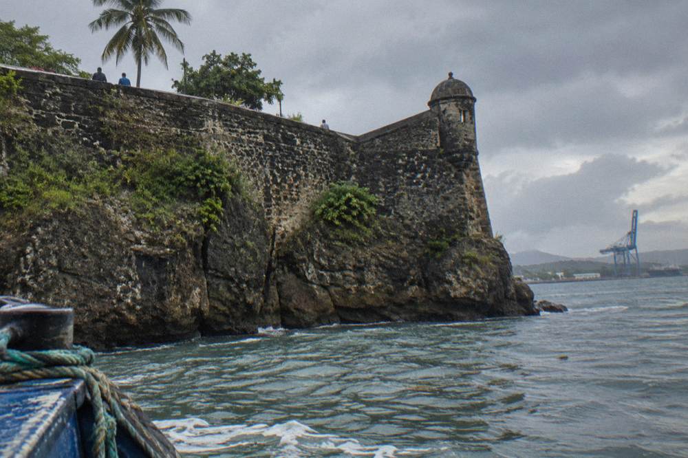 Historic stone fortress walls above a calm harbour with cargo cranes in the distance