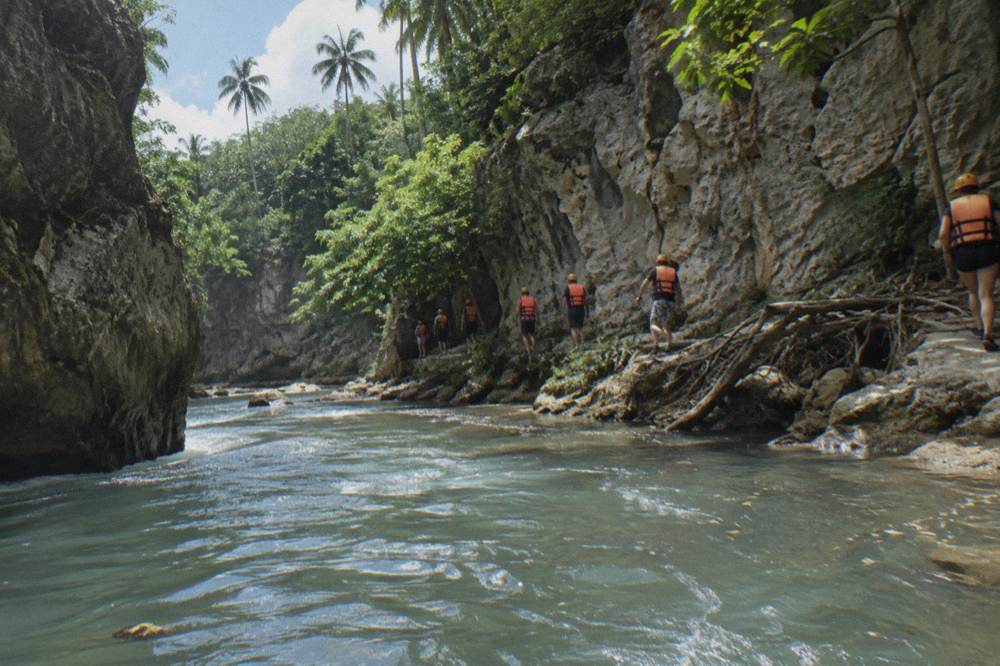 Group of helmeted visitors walking single file along a riverbank between limestone walls