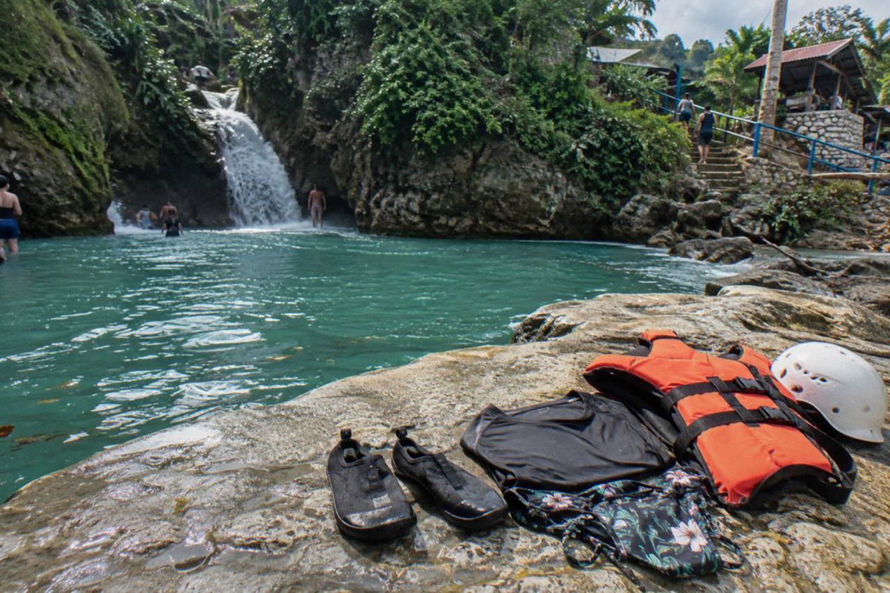Water shoes, a swimsuit, a quick-dry shirt, an orange life jacket and a white helmet on a wet limestone rock beside a turquoise pool